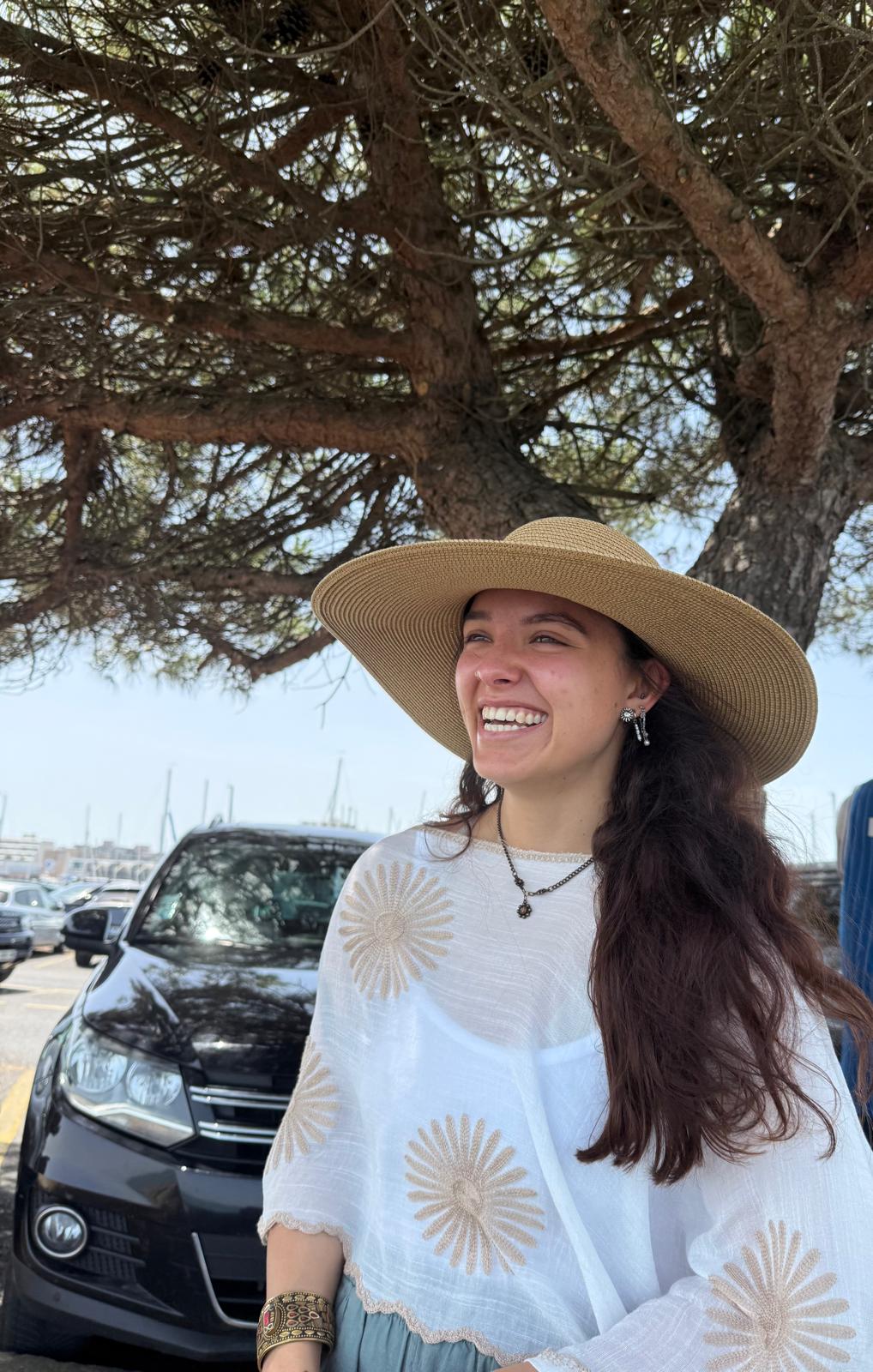 Kiki smiling beneath a tree, wearing a straw hat and a flower embroidered blouse